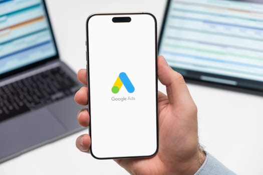 PRAGUE, CZECHIA - JULY, 2024: Google Ads logo on the screen of smartphone in mans hand and laptop on the background