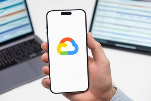 PRAGUE, CZECHIA - JULY, 2024: Google cloud logo on the screen of smartphone in mans hand and laptop on the background