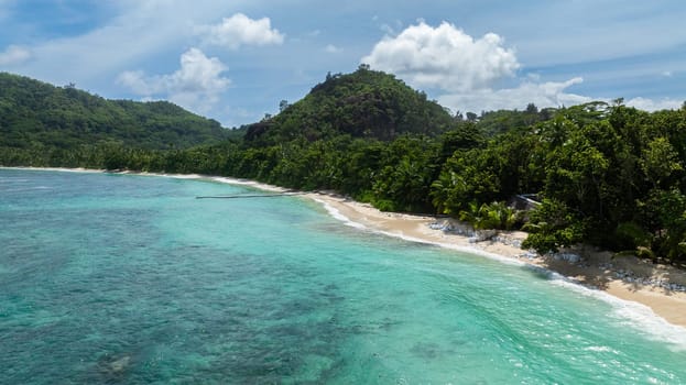 Turquoise waters gently lap against large granite rocks on a tropical beach, surrounded by lush green vegetation and palm trees. Seychelles, Mahe.