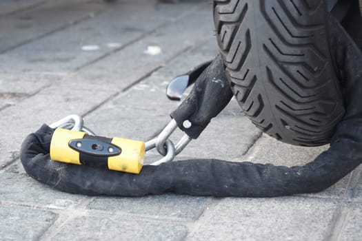 A thick cable lock connects a vehicle tire to the ground on pavement.