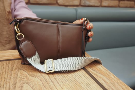 A person holds a brown handbag with a unique strap on table.
