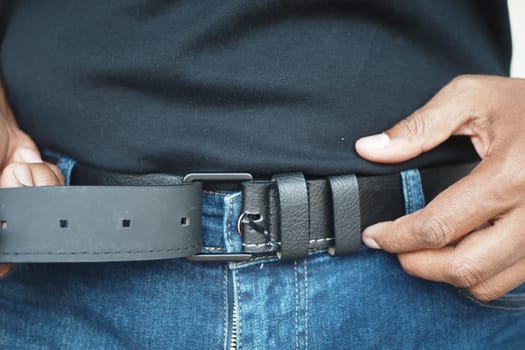 A man adjusts his black leather belt while wearing blue jeans.