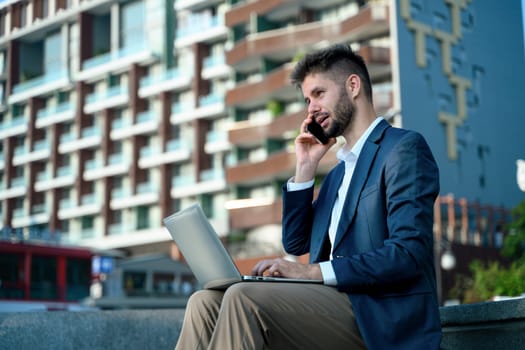 Confident businessman sitting outdoors with a laptop and speaking on his phone.