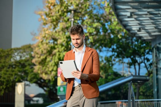 Portrait of young businessman wearing stylish suit using digital tablet outside office building.