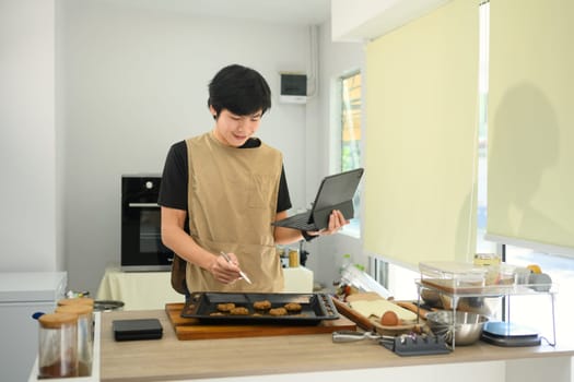 Smiling male baker checking recipe on digital tablet while preparing chocolate chip cookies.