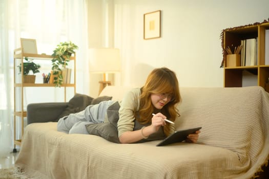 Happy young woman lying on a beige couch and drawing on a digital tablet.
