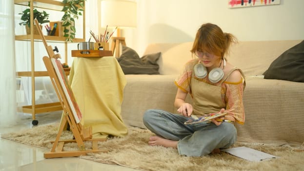 Female artist in glasses sitting cross legged on a plush rug and mixing colors on palette.