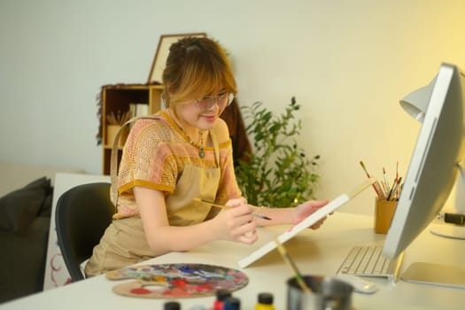 Young female artist wearing glasses painting on a canvas in a cozy home studio.