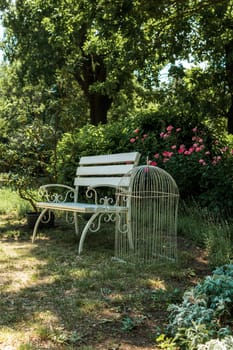 A cute white bench stands in the park among the trees. Next to it is a white iron decorative bird cage.A wooden bench in a park. High quality photo