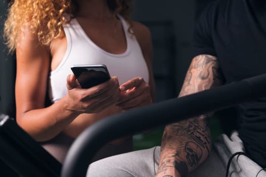 crop of unrecognizable female with curly hair in white top showing smartphone to male with tattoos in room, concept of technology of communication