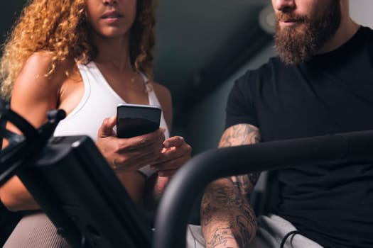 crop of unrecognizable female with curly hair in white top showing smartphone to male with tattoos in room, concept of technology of communication