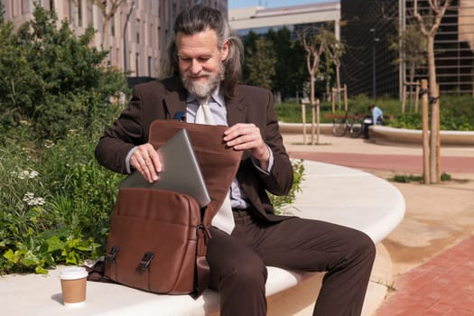 mature male lawyer in formal clothes taking laptop from briefcase while sitting with cup of takeaway coffee on urban square, concept of business and middle age man lifestyle