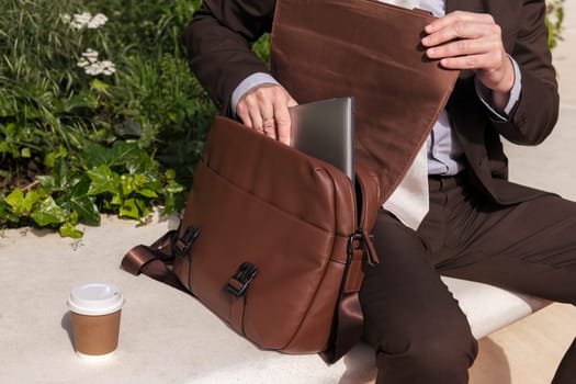 crop of unrecognizable male lawyer in formal clothes taking laptop from briefcase while sitting with cup of takeaway coffee on urban square, concept of business and urban lifestyle