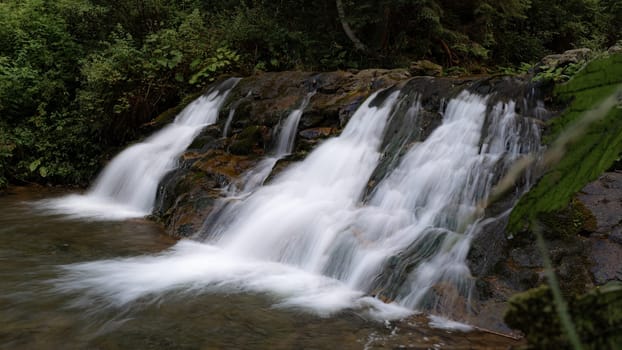 This tranquil waterfall flows gracefully over rocks, with vibrant green plants framing the scene. The soft rush of the water adds to the calming ambiance of the natural environment.