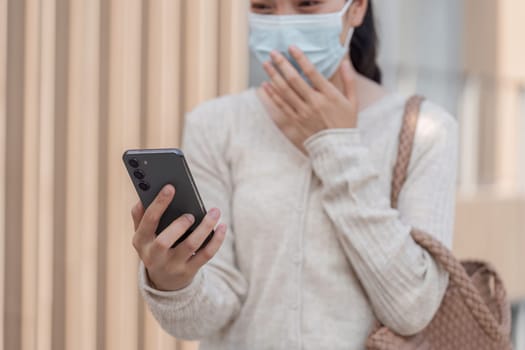 A woman wearing a mask is holding her smartphone and reacting with surprise to news while outdoors in an urban area.