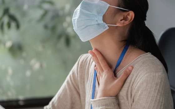 A woman wearing a mask is holding her throat while seated at her desk, suggesting discomfort or respiratory issues in a workspace.