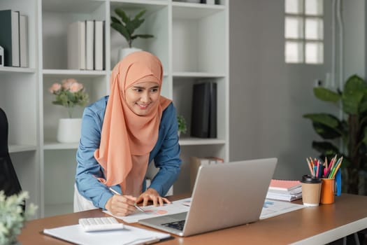 A Muslim woman wearing a hijab stands at her desk, analyzing data and making notes while looking at her laptop in a contemporary office setting.