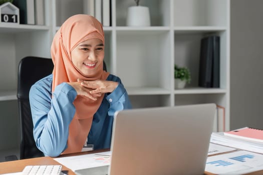 A Muslim woman in a hijab, happily engaging with her laptop and reviewing documents in a contemporary office workspace.