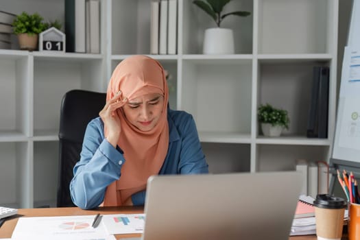A diverse Muslim woman in a hijab appearing overwhelmed and stressed while focusing on her laptop in a modern office filled with financial documents.