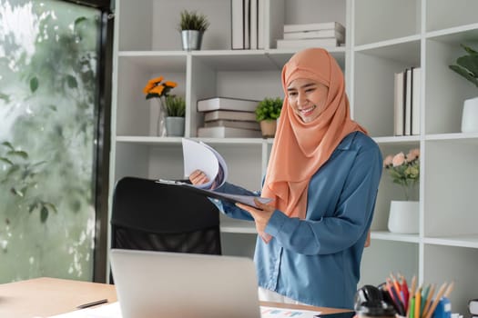 A Muslim woman in a hijab smiling as she reviews business documents in her office, surrounded by organized shelves and a laptop.