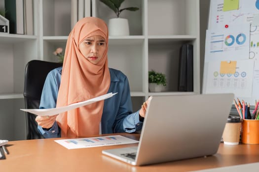 A diverse Muslim woman in a hijab looking concerned while reviewing financial documents on her laptop at her desk in a modern office environment.