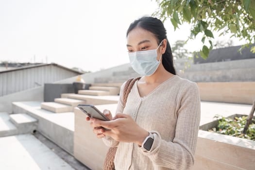 A young woman wearing a protective mask is using a smartphone in a public space, showcasing health awareness and technology use amidst outdoor settings.