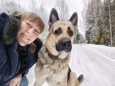 Middle-aged woman captures a fun selfie with her German shepherd during a winter camping trip in a snowy landscape.