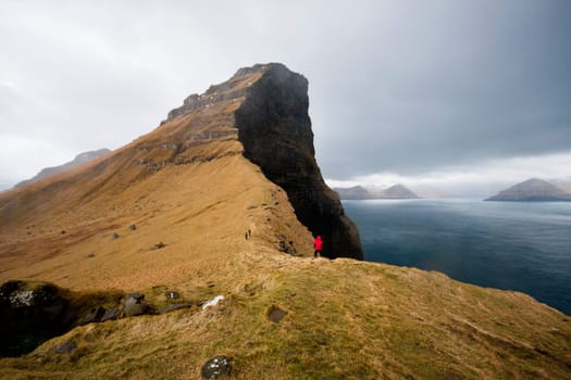Woman in a red coat exposed to the elements walking across unique and steep rock formations in stormy weather. Kalsoy Island, Faroe Islands, Denmark