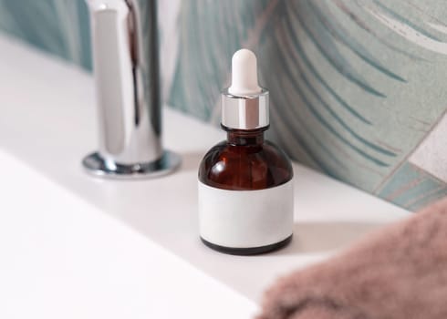 Brown glass dropper bottle with pipette near brown towels and faucet on basin in green tropical bathroom close up, label mockup. Natural unbranded cosmetics with copy space. Spa concept