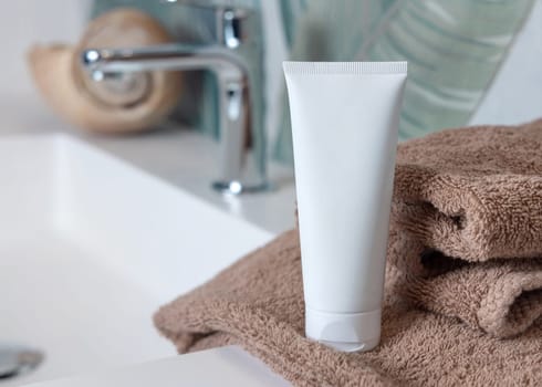 White cream squeeze tube on brown folded towels on basin in green tropical bathroom close up, packaging mockup. Natural plant unbranded cosmetics with copy space. Spa and wellness concept