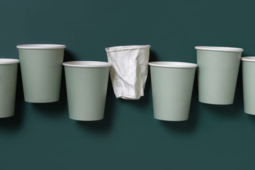 Disposable new light green paper cups and crumpled used white one in a line a dark green top view, mockup with copy space. Empty cardboard coffee cups flat lay. No plastic and sustainable lifestyle concept
