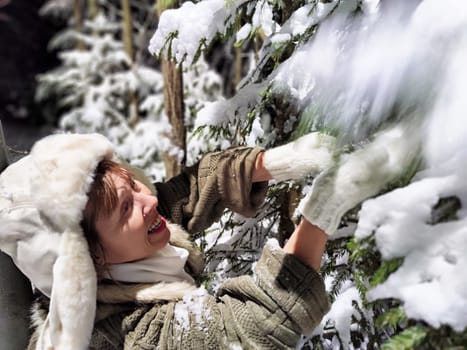 A funny middle-aged woman is cheerfully walking in the snowy forest at night, playfully interacting with the trees while wearing a unique hat.