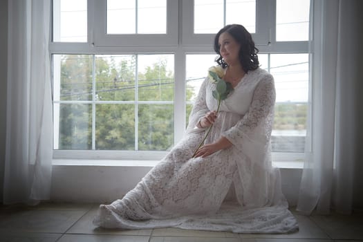 The woman in a lovely dress holds a rose while seated by a window, capturing a moment of beauty and romance in soft light.