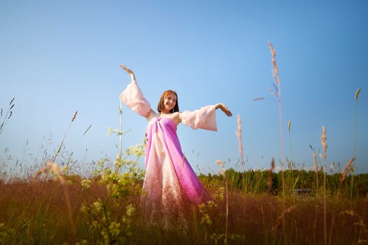 A joyful woman wearing a colorful dress dances gracefully in a sunlit meadow, surrounded by wild grasses under a clear blue sky.