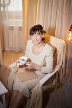 A middle-aged woman sits comfortably in her cozy apartment, savoring a warm drink while enjoying a moment of relaxation in the evening.