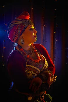 A woman in a colorful headscarf and jewelry poses in front of wall of colorful tassels