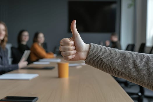 A person is giving a thumbs up in a group setting. The group is sitting around a table with various items on it, including a cup, a book, and a cell phone. Scene is positive and friendly
