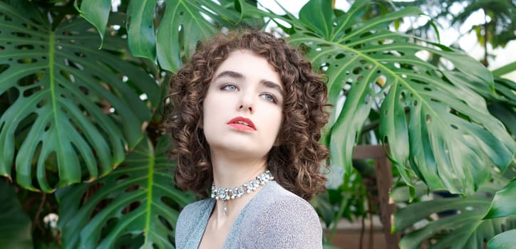 Woman with curly hair is wearing a necklace and a red lipstick. She is standing in front of a large green plant