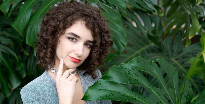 Woman with curly hair is standing in front of a green plant. She is wearing a blue dress and a necklace