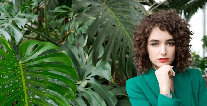 Woman with curly hair is sitting in front of a plant. She is wearing a green jacket