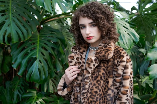 Woman wearing a leopard print coat and a necklace. She is standing in front of a green plant