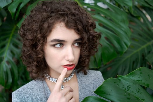 Woman with curly hair and a necklace is looking at the camera. She is wearing a gray jacket and a red necklace