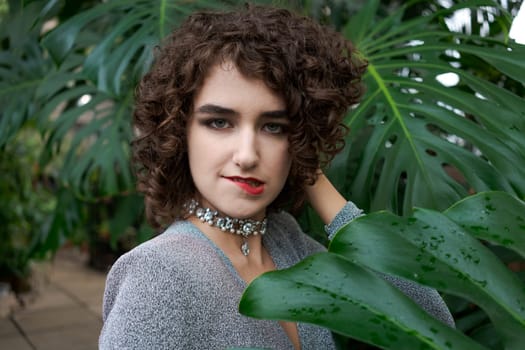 Woman with curly hair is wearing a necklace and a red lipstick. She is standing in front of a green leafy plant