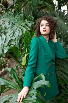 Woman in a green jacket stands in front of a plant. She is wearing a necklace and has red lipstick on