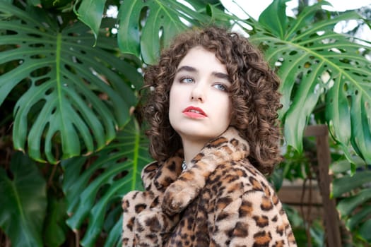 Woman with brown hair and a leopard print coat. She is standing in front of a green plant