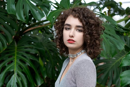 Woman with curly hair is wearing a silver necklace. She is standing in front of a green plant