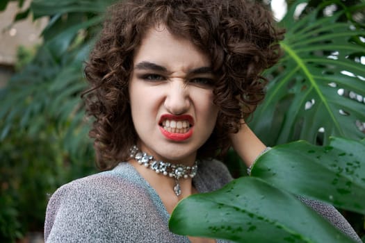 Woman with red lipstick and a necklace is holding a leaf. She has a frown on her face
