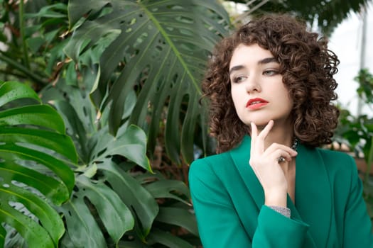 Woman with curly hair is wearing a green jacket and looking at the camera. She is wearing a necklace and a red lipstick