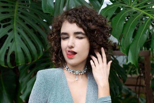 Woman with curly hair is wearing a necklace and a blue dress. She is licking her tongue