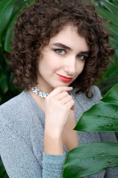 Woman with curly hair is wearing a silver necklace and a red lipstick. She is posing for a picture in front of a green plant